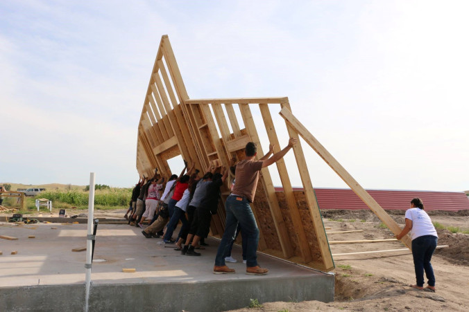 A team of people push up a frame of a wall on a cleared lot